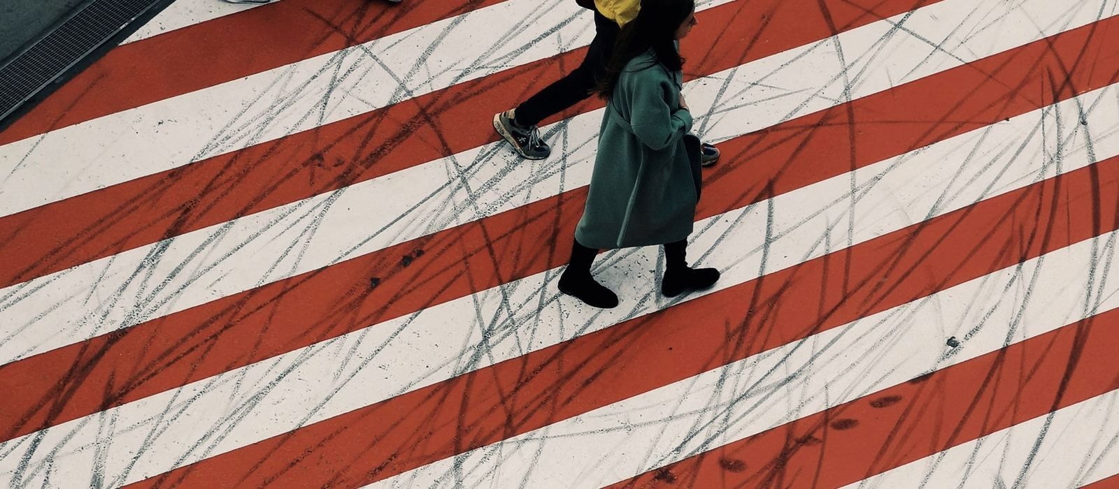 Seen from above, two people cross a road on bold painted stripes